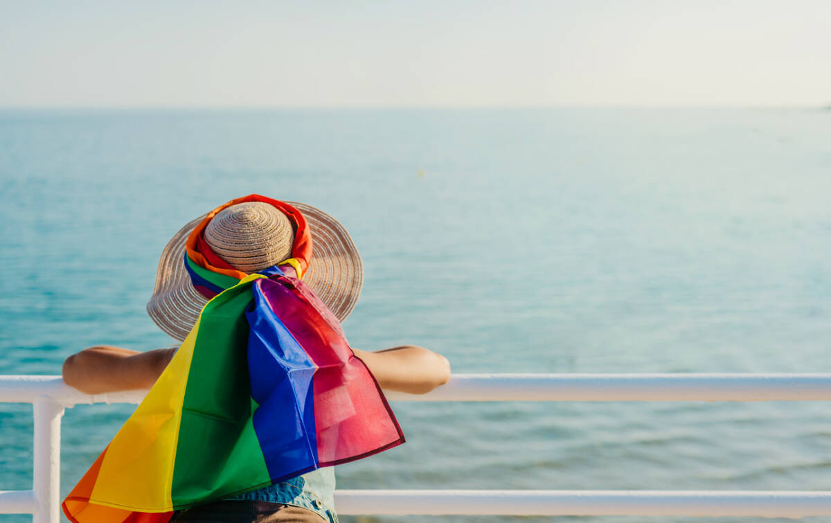 a woman leaning on a railing looks out to sea with a rainbow-colored scarf. lgtb. pride flag.