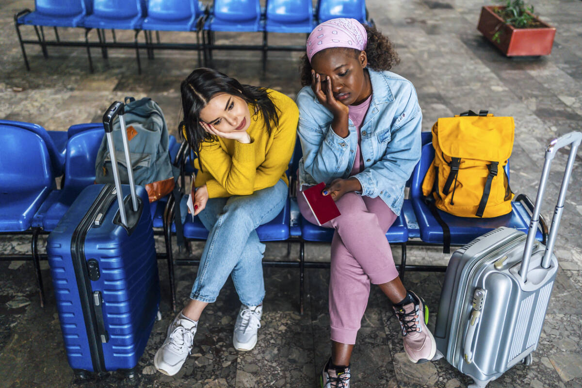 Young multi racial women friends with suitcases sitting bored, waiting for a train at railway station